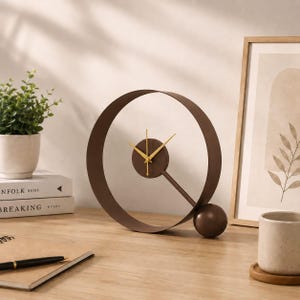 Pode incluir: Um relógio de mesa em metal castanho com uma grande moldura circular, um mostrador central com ponteiros dourados e uma pequena base esférica. O design do relógio é aberto e assenta sobre uma superfície plana com um acabamento escuro.