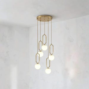May include: A multi-light pendant chandelier featuring a circular brass ceiling canopy with five hanging cords. Each cord supports an elongated oval brass frame holding a single spherical white glass globe light at varying heights.