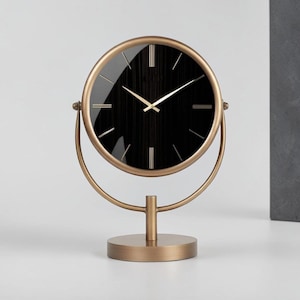 May include: A round table clock featuring a black face with a subtle vertical texture, gold-toned hands, and gold-toned hour markers. The clock is encased in a gold-toned metal frame that pivots within a circular support on a round base.
