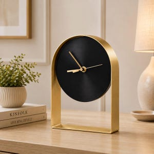 Puede incluir: Un reloj de mesa con esfera negra circular y manecillas doradas, alojado en un marco de metal dorado arqueado y liso. El reloj se apoya sobre una base rectangular, presentando un diseño metálico limpio con una superficie de esfera negra mate.