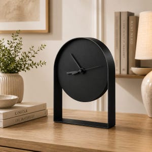 Puede incluir: Un reloj de mesa negro mate con una esfera circular y manecillas simples para las horas y los minutos, apoyado sobre una base de marco de metal arqueado. El reloj tiene un diseño limpio y de color sólido sin números ni marcas en la esfera.