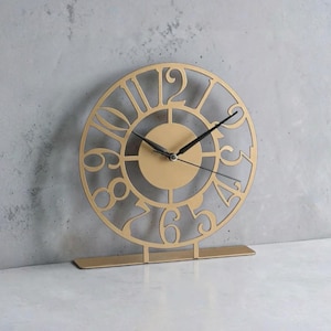 May include: A gold-colored, decorative clock with a circular face and Roman numerals. The clock has a cut-out design and black clock hands. It stands on a rectangular base, suitable for a desk or shelf. The clock measures approximately 8 inches in diameter.