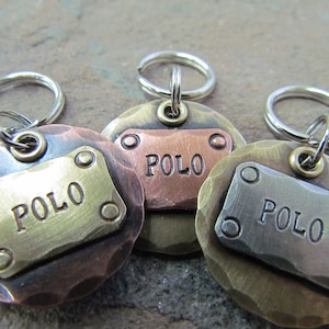 Op de afbeelding: Drie ronde, gehammerde metalen hondenpenningen met zilveren sleutelringen. Elke penning heeft een rechthoekige messingplaat met de naam "POLO" erop gegraveerd. De penningen zijn in verschillende kleuren: koper, messing en zilver.