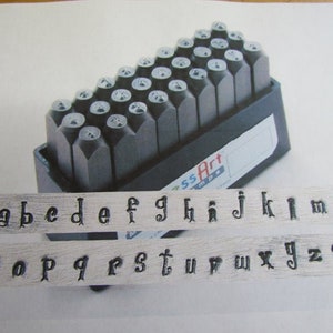 Op de afbeelding: Een set metalen letterstempels in een zwarte kunststof koffer. De stempels zijn alfabetisch gerangschikt en omvatten de letters a tot en met z, evenals de cijfers 2 en een vraagteken. De koffer heeft de tekst "ssArt mpe" erop gedrukt.