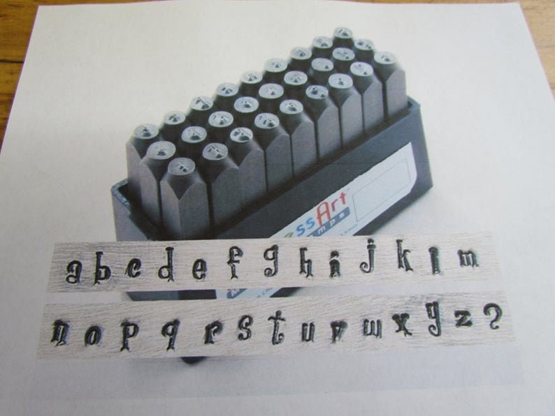 Op de afbeelding: Een set metalen letterstempels in een zwarte kunststof koffer. De stempels zijn alfabetisch gerangschikt en omvatten de letters a tot en met z, evenals de cijfers 2 en een vraagteken. De koffer heeft de tekst "ssArt mpe" erop gedrukt.