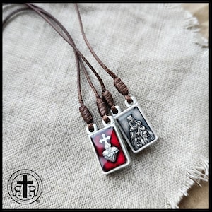May include: Two religious scapular necklaces with brown cords. One pendant features a red background with a silver heart and cross, the other depicts a religious figure. The pendants are rectangular and silver-toned.
