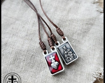 May include: Two religious scapular necklaces with brown cords. One pendant features a red background with a silver heart and cross, the other depicts a religious figure. The pendants are rectangular and silver-toned.