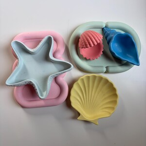 May include: A collection of silicone items including a light blue starfish-shaped bowl on a pink tray, a pale green oval tray holding a pink scallop shell and a blue conch shell, and a separate yellow scallop shell-shaped dish.