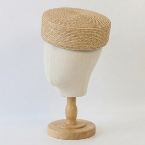 Puede incluir: Sombrero de paja beige con la parte superior plana y corona cil&iacute;ndrica. El sombrero se exhibe en una cabeza de maniqu&iacute; con un soporte de madera. La textura tejida y el color neutro sugieren un accesorio de verano.
