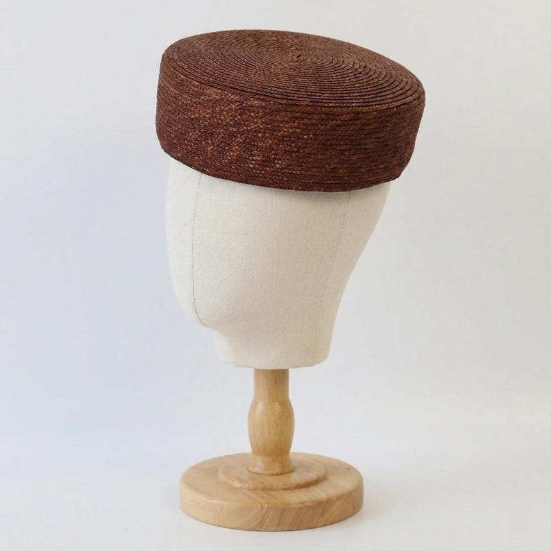 Puede incluir: Un sombrero marr&oacute;n tejido con una parte superior plana y un dise&ntilde;o circular. El sombrero se exhibe en una cabeza de maniqu&iacute; con un soporte de madera. El color del sombrero es un marr&oacute;n rico y terroso, y el tejido es apretado.