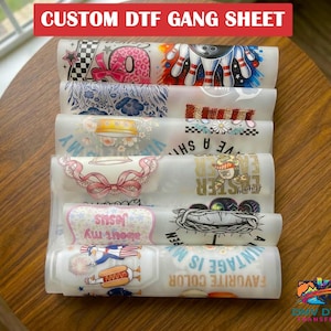 May include: A stack of printed DTF gang sheets with various designs, including a bowling pin, a pink bow, and text that reads "Jesus" and "Favorite Color." The top sheet has the text "CUSTOM DTF GANG SHEET."
