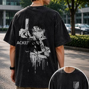 May include: A black acid-wash t-shirt featuring a large white graphic on the back of the character Levi Ackerman with the text LEVI ACKERMAN. The front of the shirt displays a small white emblem of wings and swords on the left chest area.