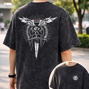 May include: Dark gray t-shirt with a large white graphic on the back featuring a winged figure, a sword, and ornate details. The shirt has a small circular design on the front. The fabric has a textured, washed appearance.