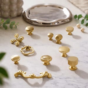 Puede incluir: Una colección de pomos y tiradores dorados para armarios en varios diseños, incluyendo estilos redondos, florales y ornamentados. Los herrajes están dispuestos sobre una superficie similar al mármol, mostrando una gama de opciones decorativas para muebles y decoración del hogar.
