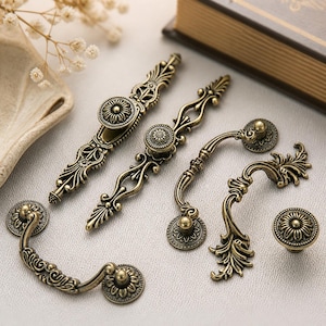 May include: An assortment of antique brass cabinet hardware, including handles and knobs. The hardware features intricate floral and scrollwork designs. The pieces vary in size and shape, with a warm, aged finish. The hardware is suitable for furniture restoration or new builds.