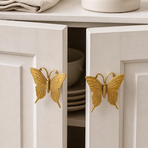 May include: Two gold-colored butterfly cabinet pulls are mounted on white cabinet doors. The butterfly pulls have detailed wing patterns and are attached to the doors with screws. The cabinet doors are slightly ajar, revealing the interior.