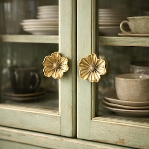 May include: Close-up of a cabinet with two gold flower-shaped knobs. The cabinet doors are painted a distressed light green color. Inside the cabinet are dishes and cups.