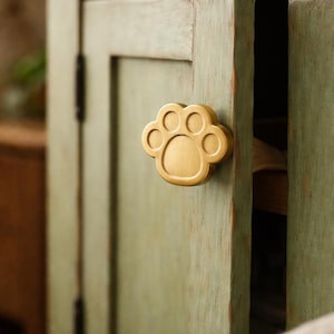 Pode incluir: Um puxador de armário dourado em forma de pegada de pata está preso a uma porta de armário verde desgastada. O puxador tem a forma de uma pata de cão ou gato, com cinco almofadas circulares. A porta do armário está ligeiramente entreaberta.