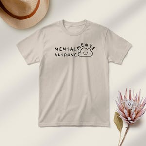 May include: A beige t-shirt with the Italian phrase "Mentalmente Altrove" printed in black, accompanied by a smiling cloud graphic. The shirt is short-sleeved and has a crew neck.