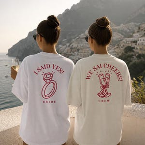Può includere: Due magliette bianche con stampe grafiche rosse sul retro. La maglietta a sinistra mostra un'icona di un anello con diamante con il testo I SAID YES! e BRIDE. Quella a destra mostra due flûte da champagne con il testo WE SAID CHEERS! e CREW.
