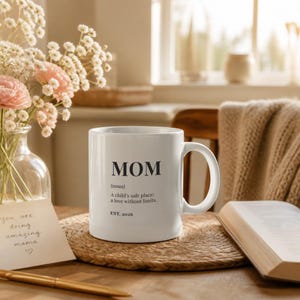Può includere: Una tazza in ceramica bianca con testo stampato in nero che recita MOM (noun) A child's safe place; a love without limits. EST. 2026. La tazza ha un manico standard e una finitura liscia, posizionata su una tovaglietta rotonda intrecciata.