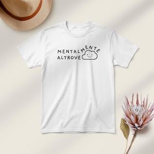 May include: White t-shirt with the Italian phrase "Mentalmente Altrove" printed in black, accompanied by a smiling cloud graphic. The shirt is short-sleeved and has a crew neck. A straw hat and a pink flower are in the background.