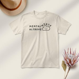 May include: Cream-colored t-shirt with the words "MENTALMENTE ALTROVE" and a smiling cloud graphic in black. A straw hat and a pink flower are in the background.