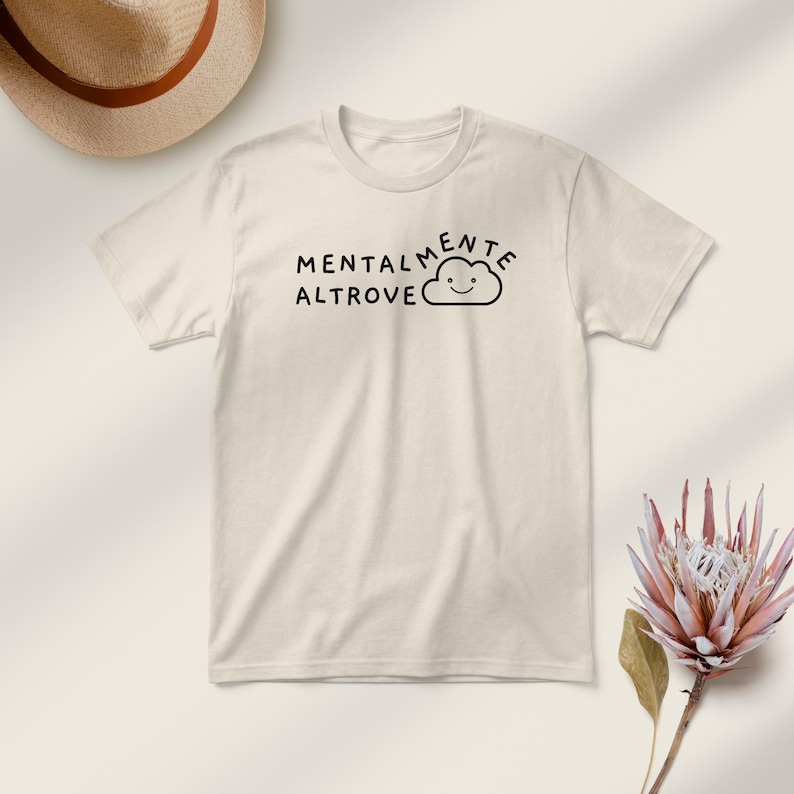 May include: Cream-colored t-shirt with the words "MENTALMENTE ALTROVE" and a smiling cloud graphic in black. A straw hat and a pink flower are in the background.