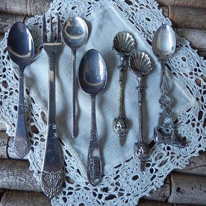 May include: An assortment of antique silver spoons and a fork are arranged on a white lace doily. The spoons have various decorative handle designs, including shell and floral motifs. The background is composed of stacked corks.