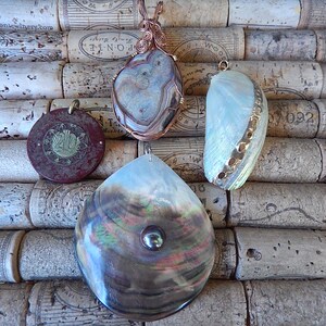 May include: An assortment of handcrafted pendants, displayed against a backdrop of corks. The pendants feature various shapes and materials, including a round, reddish-brown pendant with gold accents, and a teardrop-shaped pendant with a pearl.
