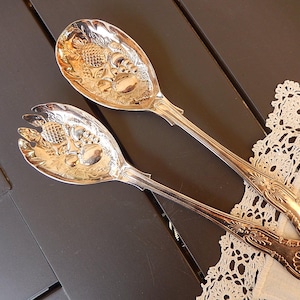 May include: Two silver serving spoons with ornate floral and fruit designs. One spoon is a serving spoon, and the other is a serving fork. The spoons are placed on a dark surface with a white lace doily.