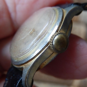 May include: Close-up of a vintage wristwatch with a gold-toned bezel and a worn, light-colored face. The watch has a black leather strap and a gold-colored crown. The watch shows signs of age and wear.