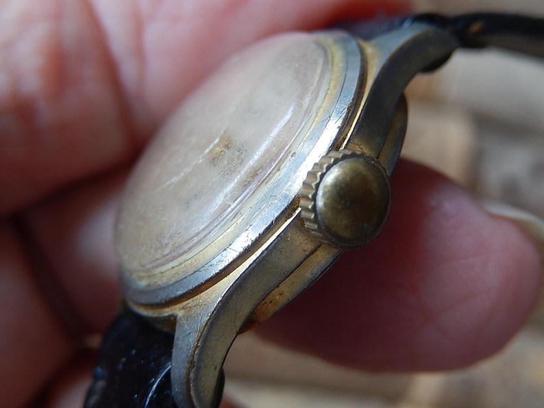 May include: Close-up of a vintage wristwatch with a gold-toned bezel and a worn, light-colored face. The watch has a black leather strap and a gold-colored crown. The watch shows signs of age and wear.