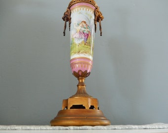 May include: A tall, ornate porcelain vase with a pink and gold base. The vase features a painted scene of a couple in a garden setting. The vase is topped with a gold metal lid and has decorative gold accents.