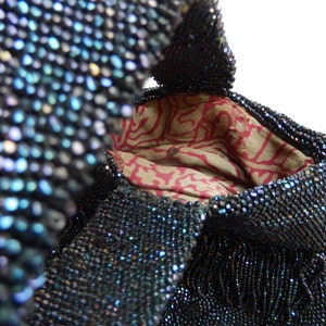 May include: A vintage beaded bag with a dark blue iridescent finish. The bag has a drawstring closure and a fringed bottom. The interior lining is a red and white floral pattern.