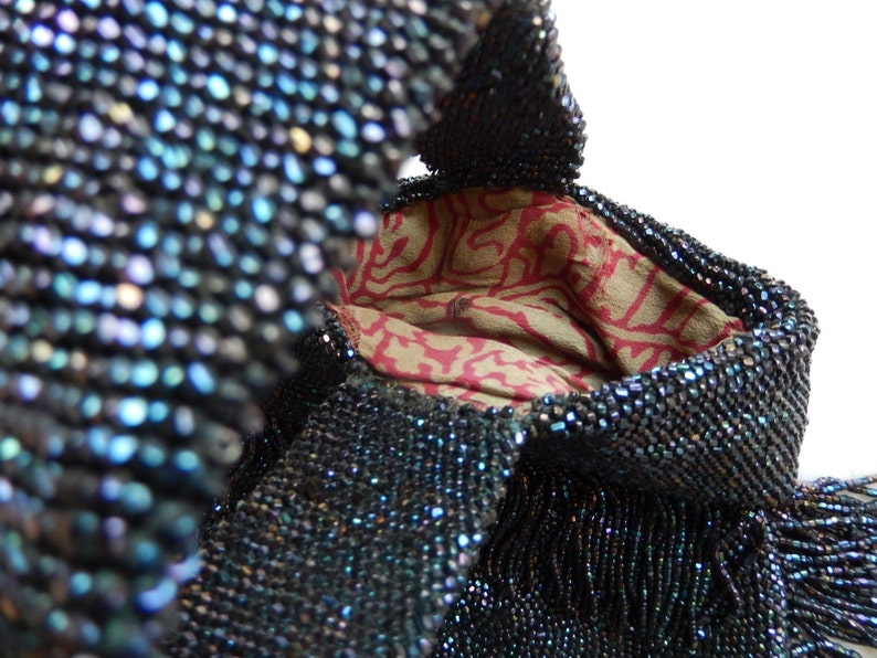 May include: A vintage beaded bag with a dark blue iridescent finish. The bag has a drawstring closure and a fringed bottom. The interior lining is a red and white floral pattern.