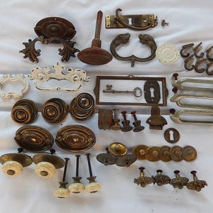 May include: A collection of antique hardware pieces, including drawer pulls, knobs, and keyhole covers. The hardware features various designs, including ornate floral patterns and simple, rounded shapes. The colors range from antique brass to white and dark brown.