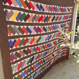 May include: A colorful quilt with a brown border and a repeating pattern of diagonal fabric pieces in various colors. The quilt is hanging outdoors, showcasing its handmade design and vibrant hues.