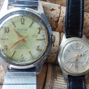 May include: Two vintage wristwatches. The larger watch has a silver-tone case and a light green face with gold-tone hands and numerals. The smaller watch has a silver-tone case and a white face with black numerals and a black leather strap.