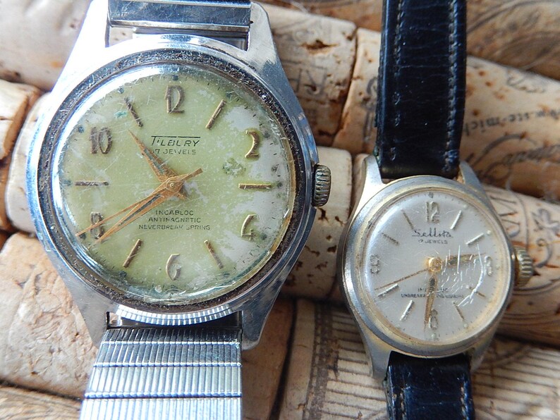May include: Two vintage wristwatches. The larger watch has a silver-tone case and a light green face with gold-tone hands and numerals. The smaller watch has a silver-tone case and a white face with black numerals and a black leather strap.