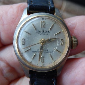 May include: Vintage Sellita wristwatch with a silver face, gold hands, and a black leather band. The watch face displays the text "Sellita 17 Jewels" and "IN BLOC UNBREAKABLE MAINSPRING". The watch has a gold-colored bezel and crown.