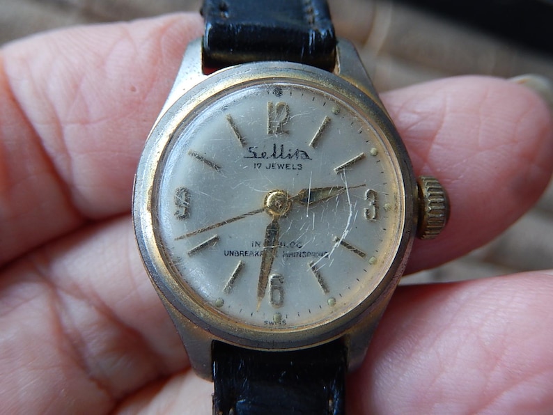 May include: Vintage Sellita wristwatch with a silver face, gold hands, and a black leather band. The watch face displays the text "Sellita 17 Jewels" and "IN BLOC UNBREAKABLE MAINSPRING". The watch has a gold-colored bezel and crown.