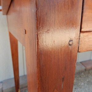 May include: Close-up of a wooden table leg with a dark brown stain. The leg is made of solid wood and has a smooth finish. The table leg is part of a wooden table with a drawer.