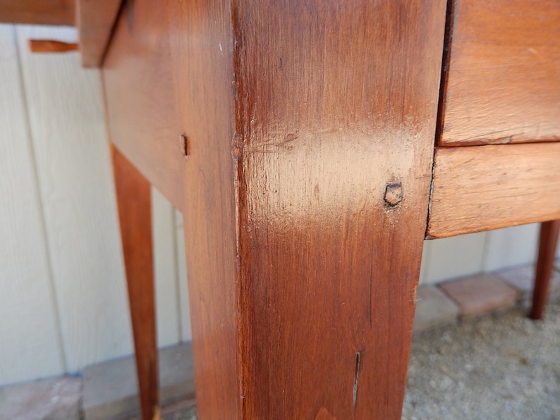 May include: Close-up of a wooden table leg with a dark brown stain. The leg is made of solid wood and has a smooth finish. The table leg is part of a wooden table with a drawer.