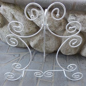 May include: White metal decorative stand with a scrolled design. The stand has a flat top and a base with three legs.
