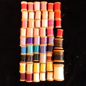 Pu&ograve; includere: Una collezione di bobine di filo colorate, disposte in file, con bobine di legno. Le bobine sono in varie tonalit&agrave; di rosso, rosa, viola, blu, verde, giallo e marrone.