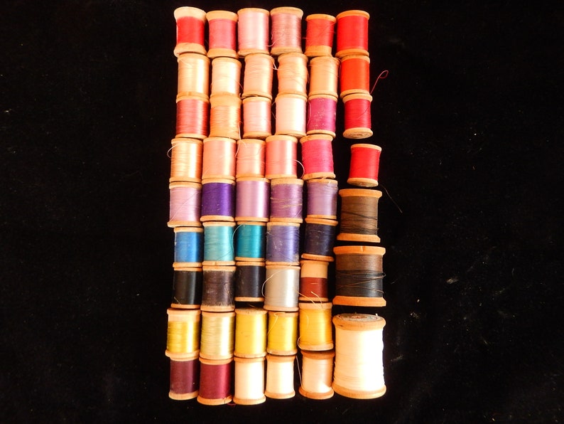 Pu&ograve; includere: Una collezione di bobine di filo colorate, disposte in file, con bobine di legno. Le bobine sono in varie tonalit&agrave; di rosso, rosa, viola, blu, verde, giallo e marrone.
