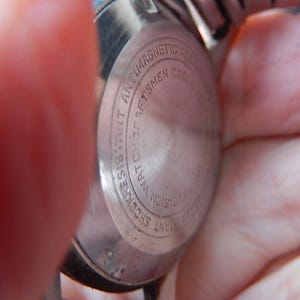 May include: Close-up of the back of a silver wristwatch. The watch casing is circular with the words "Shock Resistant," "Antimagnetic," and "Watchcraftsmen" engraved around the edge. The watch is held by a person.