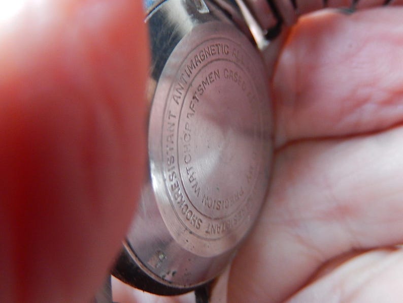 May include: Close-up of the back of a silver wristwatch. The watch casing is circular with the words "Shock Resistant," "Antimagnetic," and "Watchcraftsmen" engraved around the edge. The watch is held by a person.