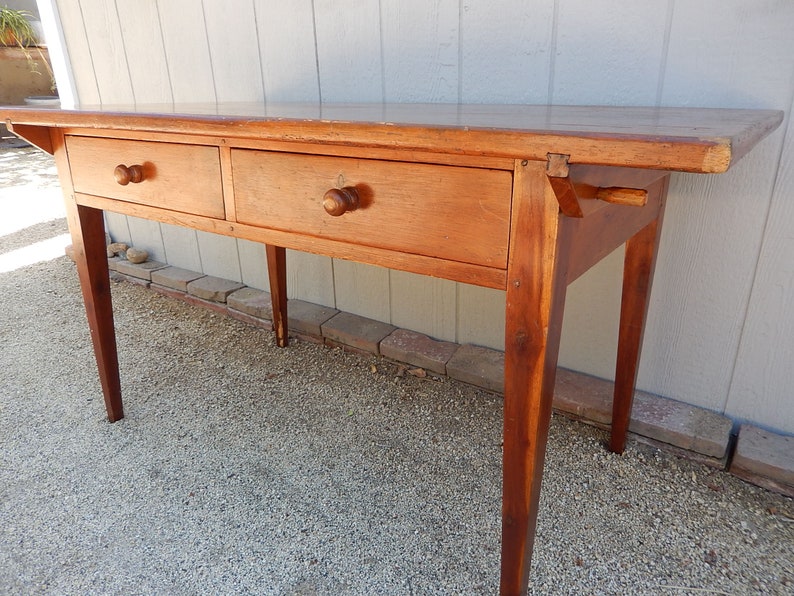 May include: A long, wooden table with two drawers and turned legs. The table is made of a light brown wood and has a simple, rustic design.